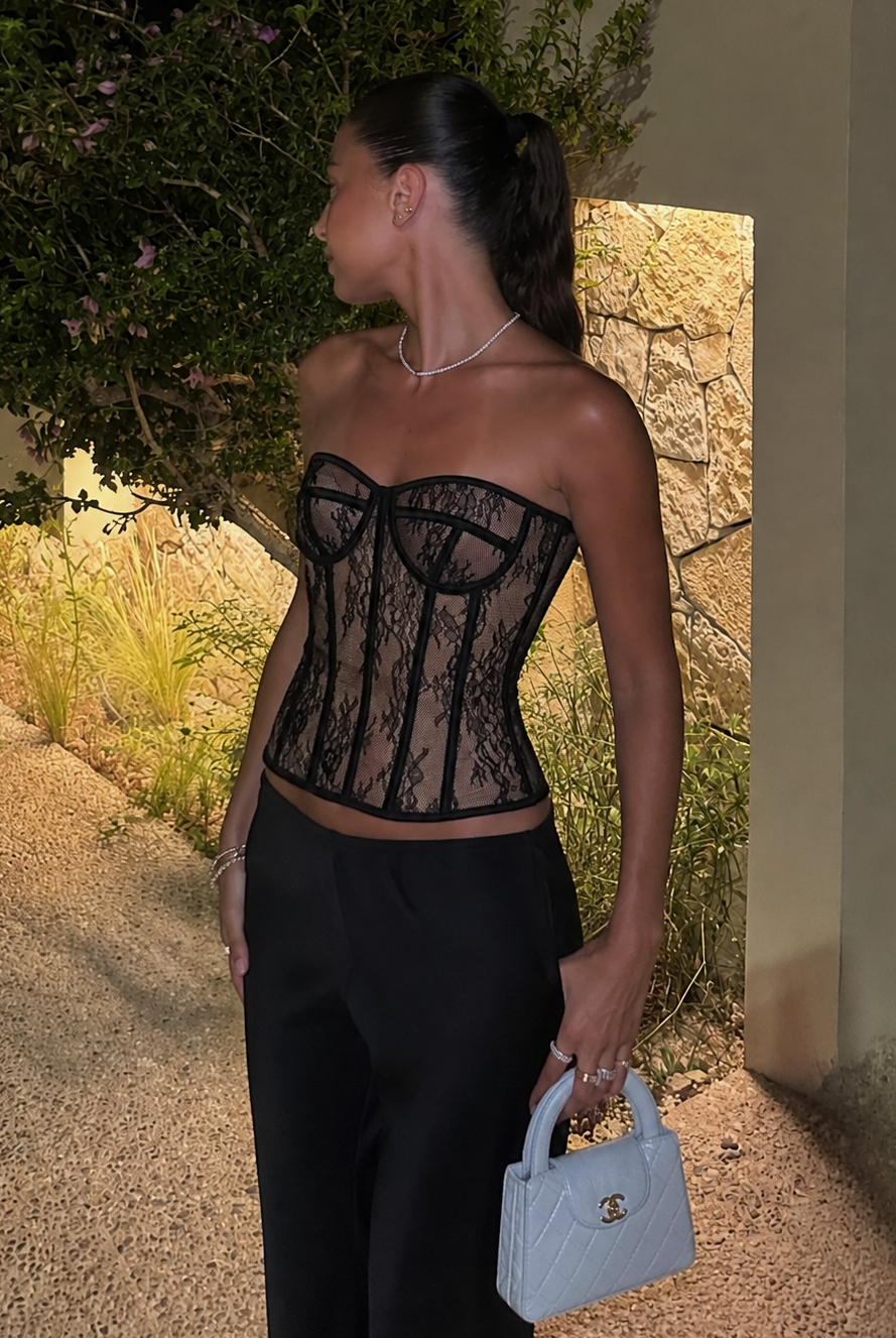A woman stands outdoors at night wearing a black lace corset top and black pants, holding a small light blue handbag. The setting includes a stone wall, plants, and warm lighting.