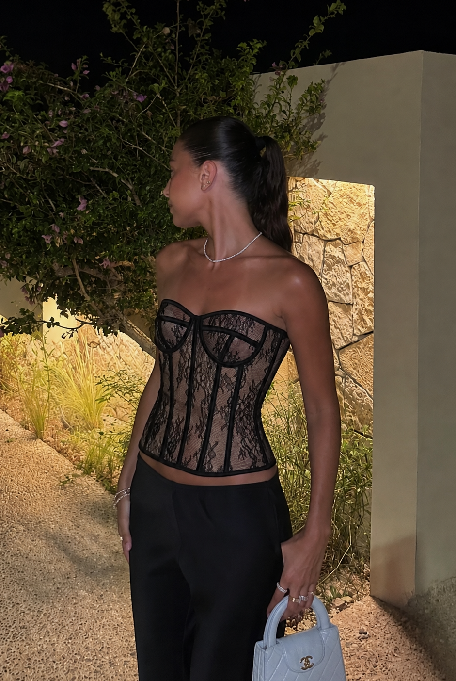 A woman stands outdoors at night wearing a black lace corset top and black pants, holding a small light blue handbag. The setting includes a stone wall, plants, and warm lighting.