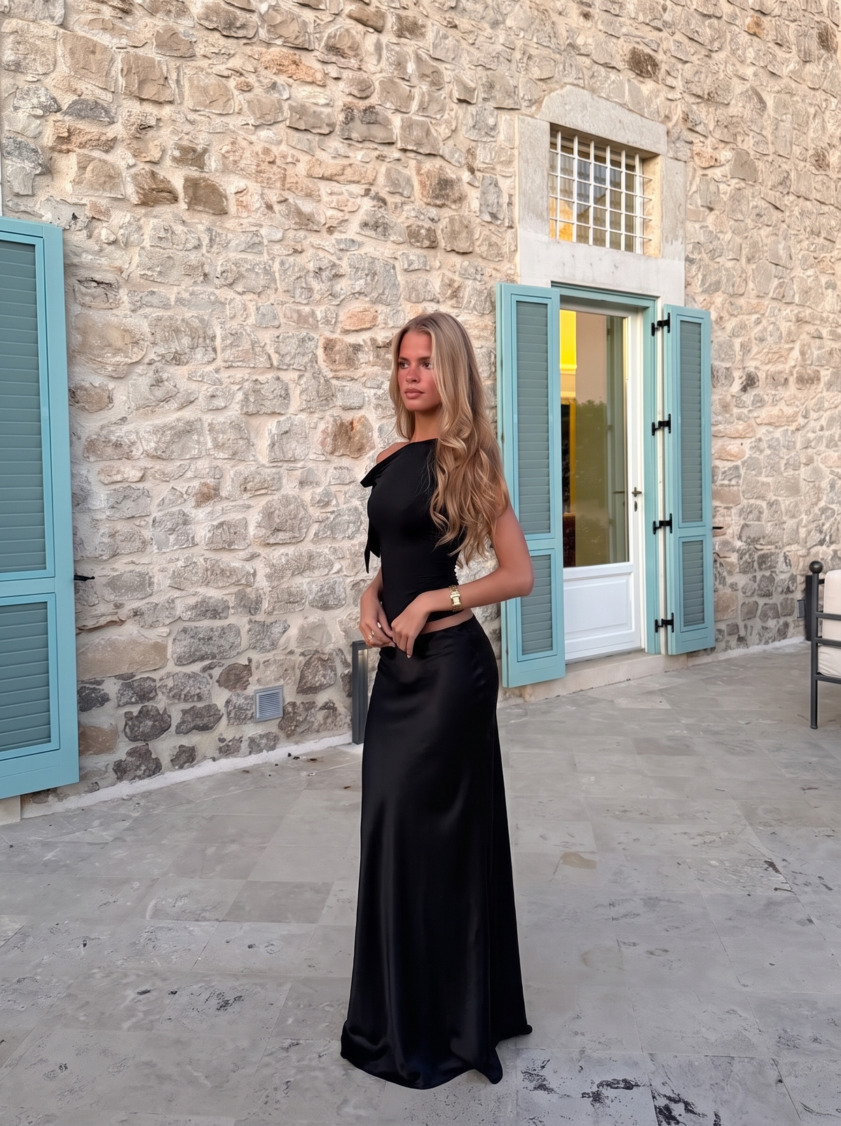 A woman wearing a black satin evening gown with a one-shoulder neckline and a fitted silhouette, standing outdoors in front of a stone building with light blue shutters.