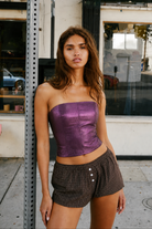 A woman stands on a city sidewalk wearing a metallic purple strapless tube top and brown plaid shorts with white buttons.