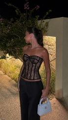 A woman stands outdoors at night wearing a black lace corset top and black pants, holding a small light blue handbag. The setting includes a stone wall, plants, and warm lighting.