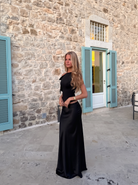 A woman wearing a black satin evening gown with a one-shoulder neckline and a fitted silhouette, standing outdoors in front of a stone building with light blue shutters.