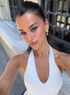 A woman taking a selfie outdoors wearing a white halter-neck top and gold drop earrings, with a stone wall and metal gate in the background.