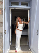 A woman models a white two-piece outfit consisting of a sleeveless crop top and a matching long skirt, standing in an open doorway of a light blue house.